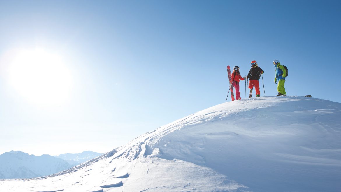 Vier Skifahrer stehen auf einer verschneiten Bergkuppe bei strahlend blauem Himmel und Sonnenschein.