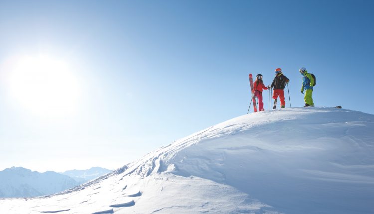 Vier Skifahrer stehen auf einer verschneiten Bergkuppe bei strahlend blauem Himmel und Sonnenschein.