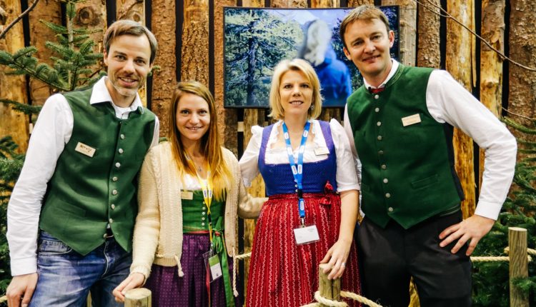 Vier Personen in traditioneller österreichischer Tracht stehen lächelnd vor einer rustikalen Holzkulisse zusammen.