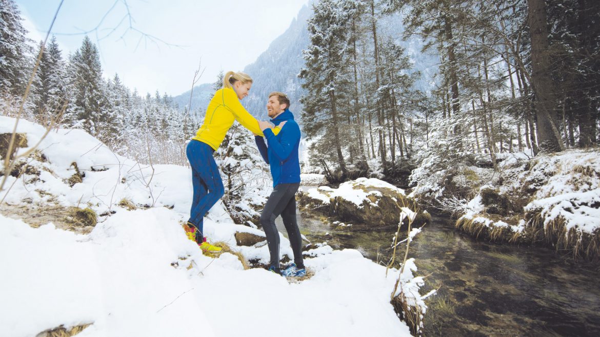 Zwei Personen in hellen Jacken stehen auf verschneitem Boden an einem Bach in einem Winterwald und lächeln sich an.