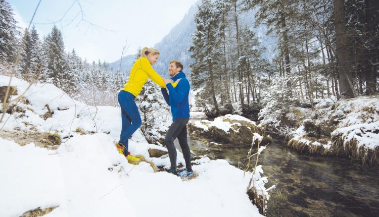 Zwei Personen in hellen Jacken stehen auf verschneitem Boden an einem Bach in einem Winterwald und lächeln sich an.