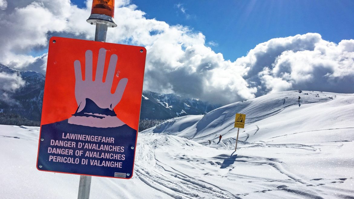 Warnschild mit roter Hand und Lawinengefahr auf verschneitem Berg bei teilweise bewölktem Himmel.