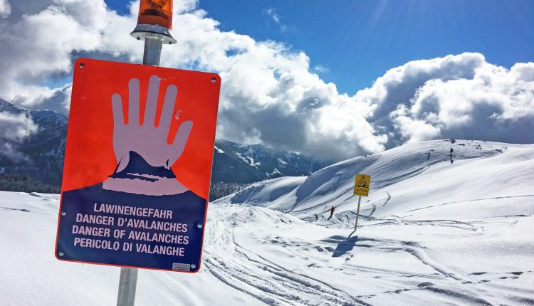 Warnschild mit roter Hand und Lawinengefahr auf verschneitem Berg bei teilweise bewölktem Himmel.