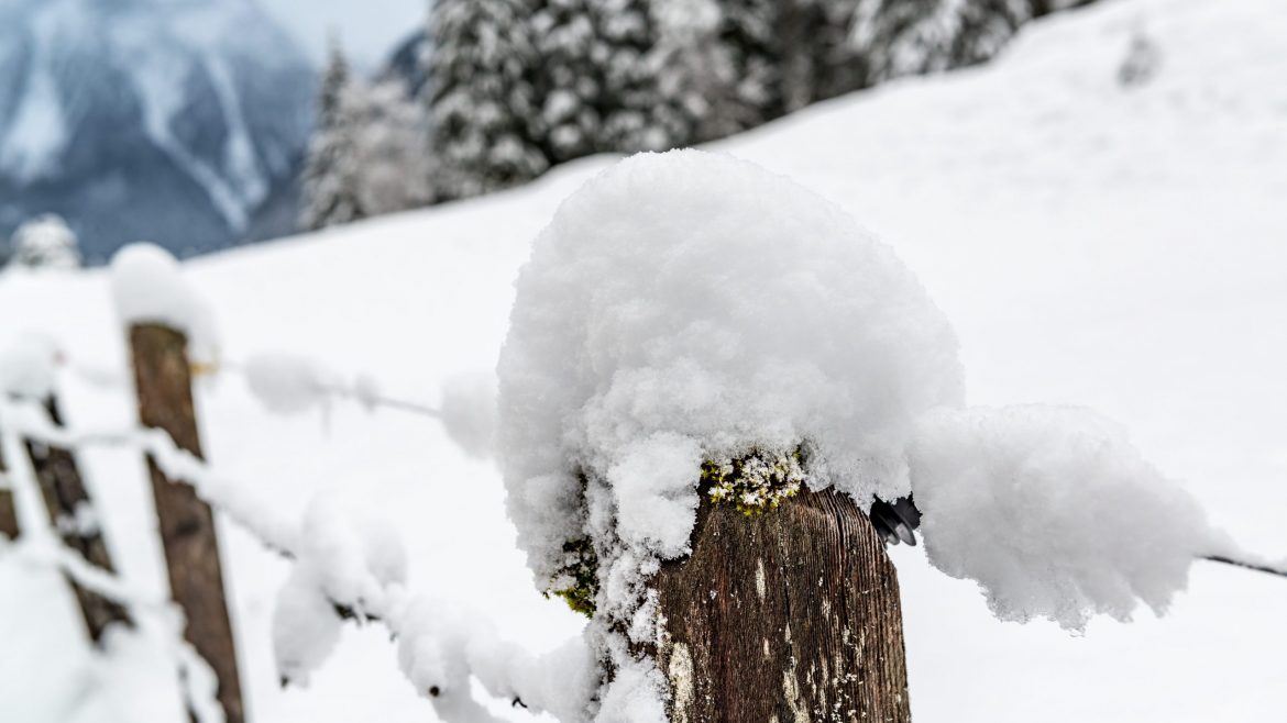 Ein hölzerner Zaunpfahl, bedeckt mit dichtem Schnee, mit verschneiten Bäumen und Bergen im Hintergrund.