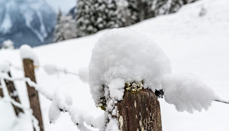 Ein hölzerner Zaunpfahl, bedeckt mit dichtem Schnee, mit verschneiten Bäumen und Bergen im Hintergrund.
