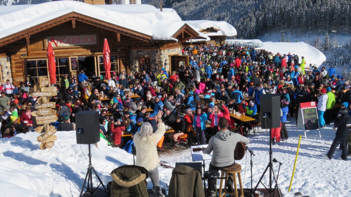 Eine große Menschenmenge in Winterkleidung versammelt sich vor einer verschneiten Berghütte zu einer Veranstaltung oder Party im Freien.