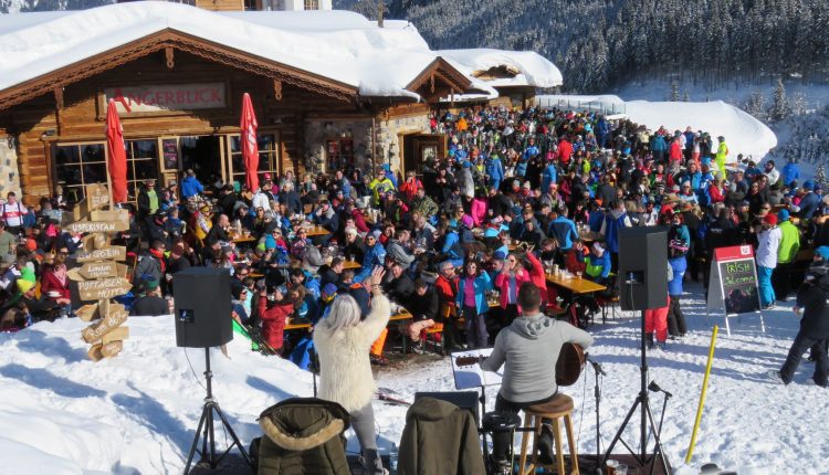 Eine große Menschenmenge in Winterkleidung versammelt sich vor einer verschneiten Berghütte zu einer Veranstaltung oder Party im Freien.