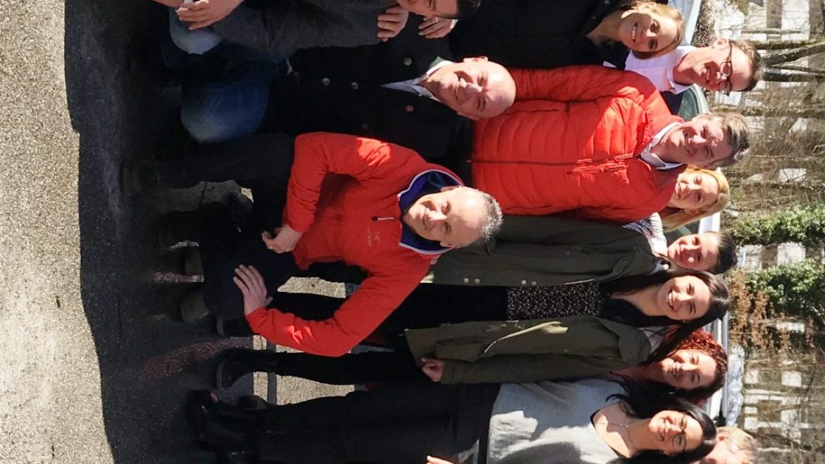 Eine Gruppe von Menschen posiert an einem sonnigen Tag im Freien, sie stehen und knien in Reihen und lächeln in die Kamera.