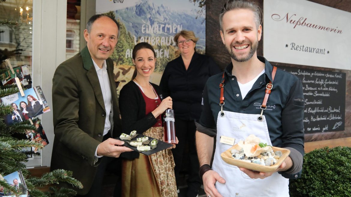 Vier Personen, die lächelnd Käseproben vor einem Restaurant mit alpenländischem Dekor servieren.
