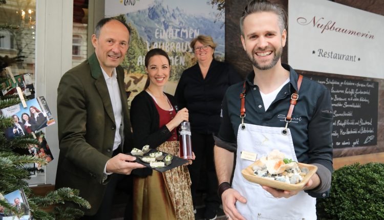 Vier Personen, die lächelnd Käseproben vor einem Restaurant mit alpenländischem Dekor servieren.