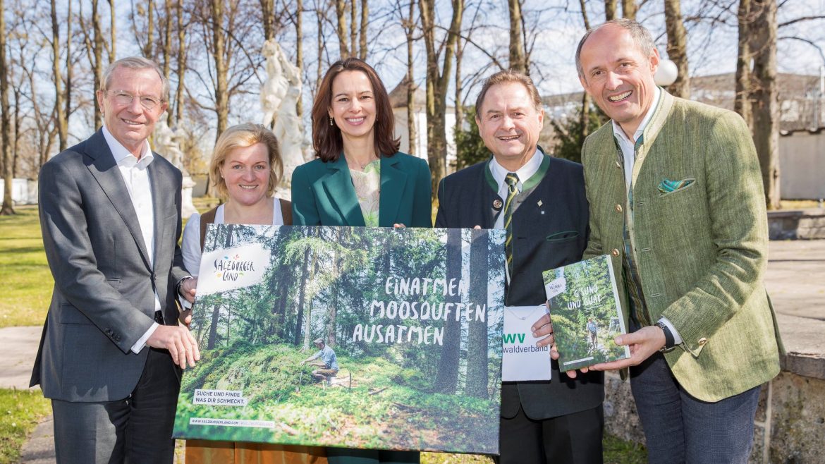Fünf Personen im Freien, die lächelnd ein großes Poster mit einer Waldszene und einem Buch in der Hand halten, mit Bäumen im Hintergrund.