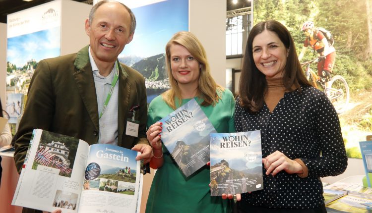 Drei Personen halten an einem Stand auf einer Tourismusmesse Reisemagazine und Broschüren hoch, im Hintergrund sind Bilder von Bergen zu sehen.