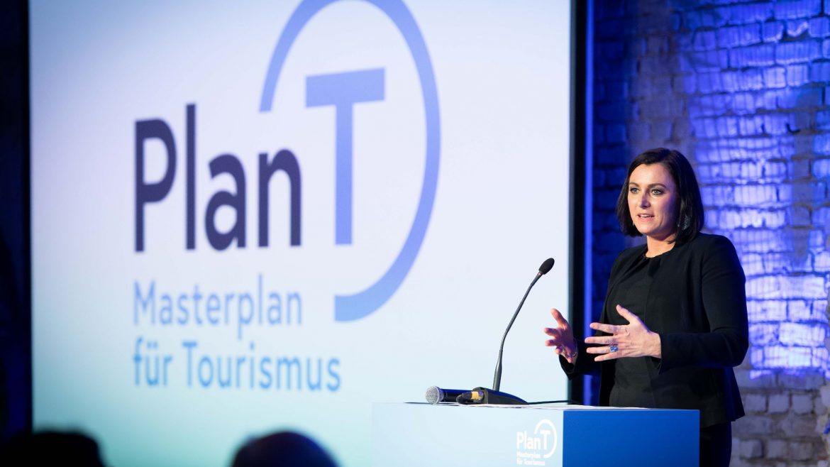 Eine Frau spricht an einem Podium mit einem Plan T Masterplan für Tourismus Schild und Logo im Hintergrund.