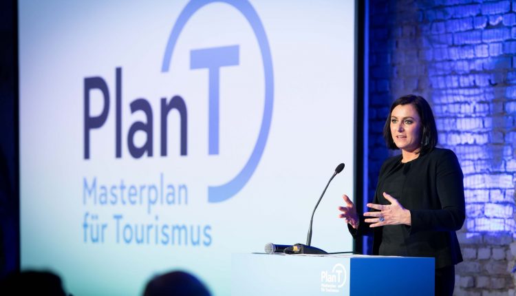 Eine Frau spricht an einem Podium mit einem Plan T Masterplan für Tourismus Schild und Logo im Hintergrund.