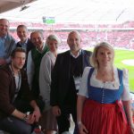 Eine Gruppe lächelnder Menschen in einer Stadionsuite, im Hintergrund ist ein Fußballfeld zu sehen.