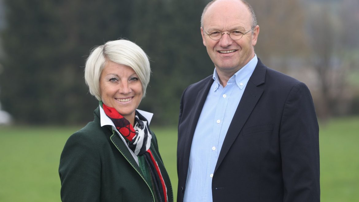 Eine lächelnde Frau und ein lächelnder Mann stehen zusammen im Freien mit Grün und Bäumen im Hintergrund.