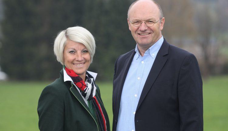 Eine lächelnde Frau und ein lächelnder Mann stehen zusammen im Freien mit Grün und Bäumen im Hintergrund.