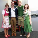 Vier lächelnde Personen, zwei davon in traditioneller österreichischer Kleidung, stehen in einem Bürogebäude.