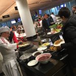 Ein Koch bereitet das Essen an einem Buffet zu, während sich die Gäste in einem belebten Essbereich anstellen und bedienen.