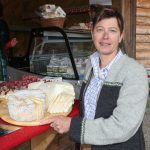 Frau präsentiert verschiedene Käsesorten auf einer Holzplatte an einem Marktstand.