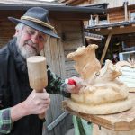 Ein älterer Mann mit Hut schnitzt im Freien mit Hammer und Meißel eine Holzskulptur und lächelt in die Kamera.