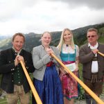 Vier Personen in traditioneller österreichischer Kleidung, die Alphörner in der Hand halten, stehen im Freien in einer bergigen Gegend.