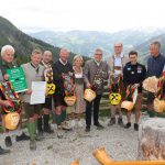 Eine Gruppe von Menschen in traditioneller österreichischer Alpenkleidung hält geschmückte Kuhglocken im Freien mit Bergen im Hintergrund.