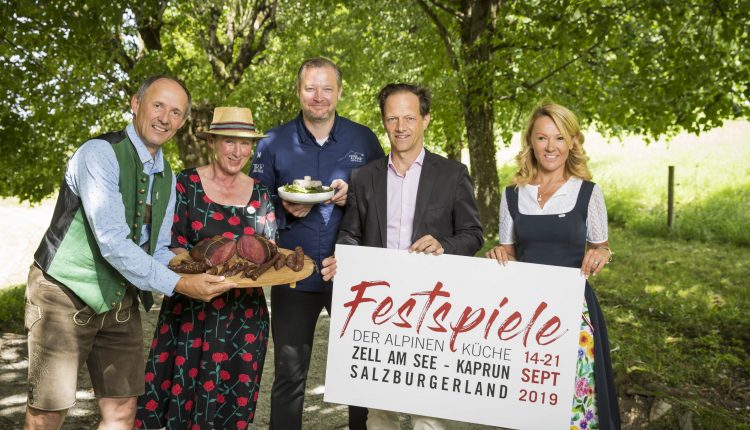 Fünf Personen in österreichischer Tracht halten an einem sonnigen Tag im Freien unter Bäumen Essen und ein Festschild in der Hand.