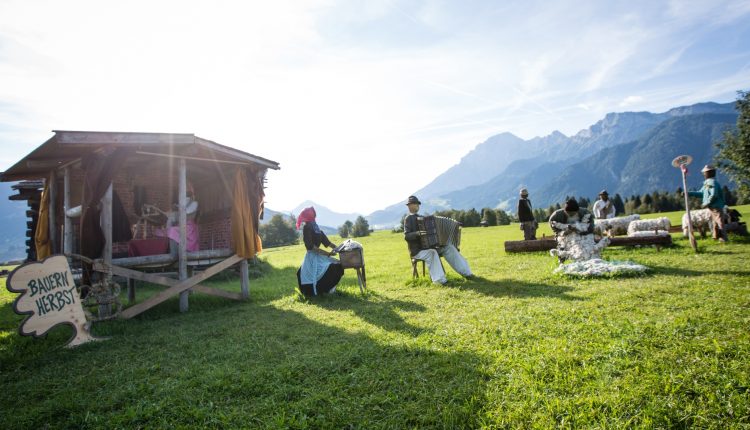 Menschen in traditioneller Kleidung sitzen neben einer Holzhütte auf einer sonnigen Bergwiese mit einem Bauernherbst-Schild.