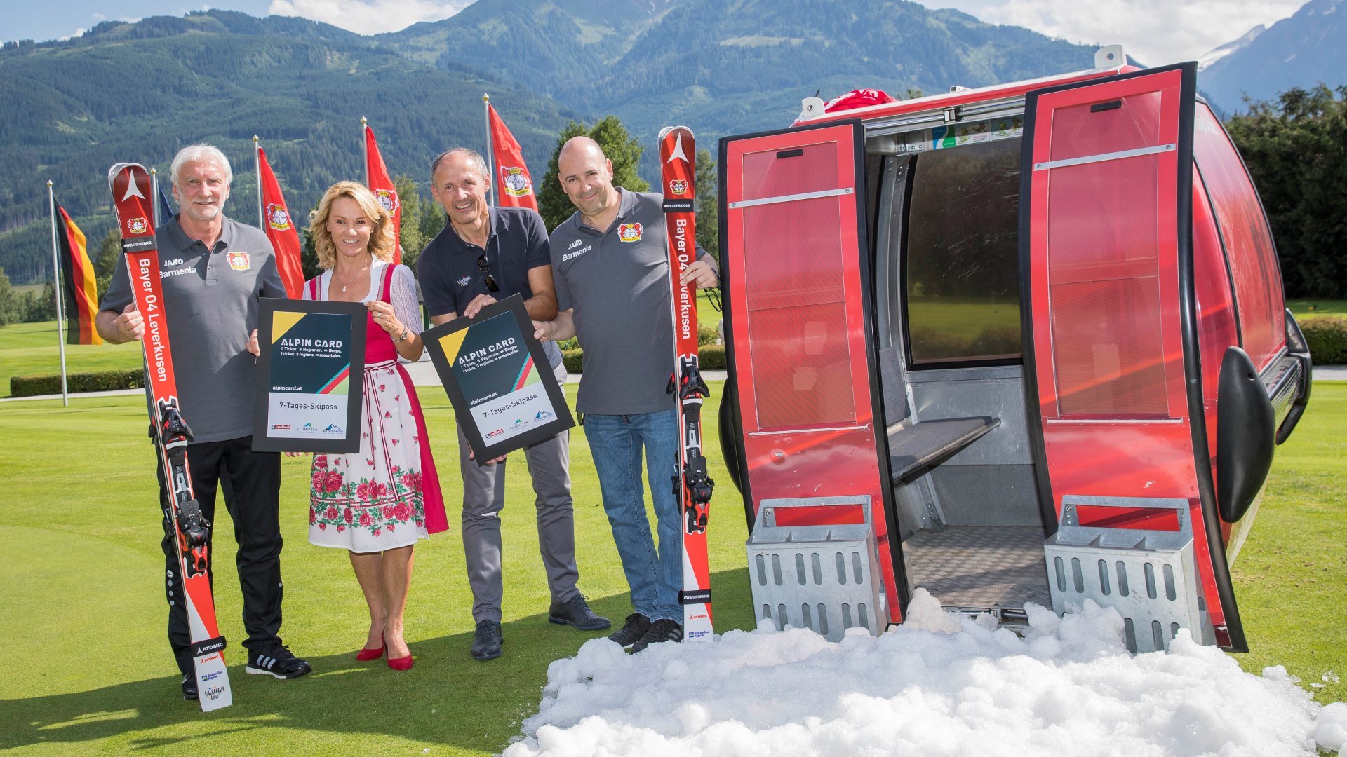 Vier Personen mit Schildern und Skiern stehen im Gras neben einer Skigondel, im Hintergrund die Berge.