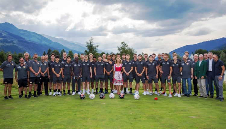 Eine große Fußballmannschaft posiert im Freien auf einer Wiese mit Bergen im Hintergrund, in der Mitte steht eine Frau in einem Kleid.