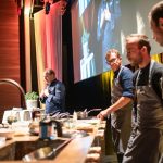 Vier Köche geben in einer Küche auf der Bühne eine Kochvorführung vor Publikum.