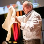 Ein Koch dehnt Pizzateig in einer Küche aus, mit bunten Lichtern und einem Bildschirm im Hintergrund.