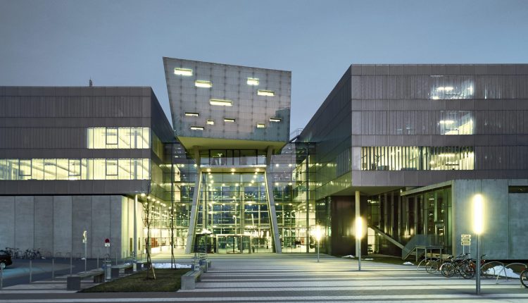 Modernes Gebäude aus Glas und Beton mit geometrischer Architektur, das in der Abenddämmerung beleuchtet wird, vor dem Fahrräder geparkt sind.