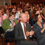 Publikum in formeller Kleidung sitzt und applaudiert bei einer Veranstaltung oder Konferenz in einem Gebäude.