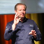 Ein Chefkoch in einer blauen Jacke gestikuliert mit den Händen, während er auf der Bühne spricht.