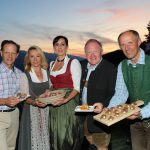 Fünf Personen in traditioneller alpiner Kleidung halten Tabletts mit Vorspeisen im Freien bei Sonnenuntergang und lächeln in die Kamera.