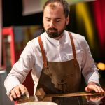 Ein Koch in einer braunen Schürze bereitet unter Bühnenbeleuchtung auf einem Tresen Speisen zu.