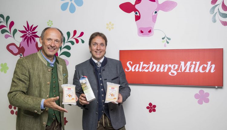 Zwei Männer in traditioneller österreichischer Kleidung halten SalzburgMilch-Milchprodukte in der Hand und stehen neben einem roten SalzburgMilch-Schild.