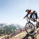 Mountainbiker in voller Montur rast bergab über Felsen, mit einer Menschenmenge und Bergen im Hintergrund.