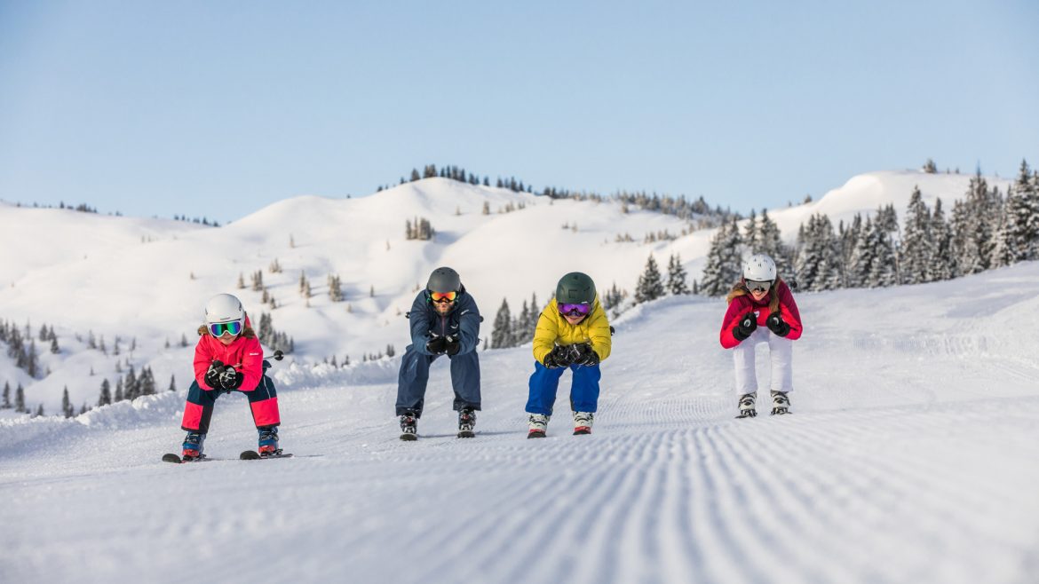 Vier Kinder in bunten Skiklamotten fahren auf einem sonnigen, verschneiten Berg mit Bäumen im Hintergrund bergab.