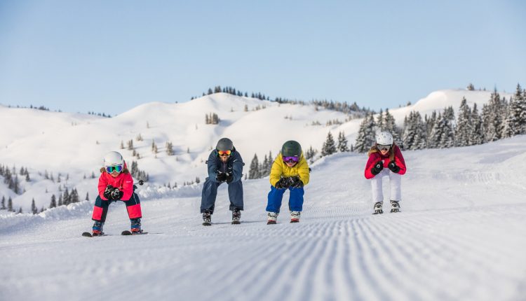 Vier Kinder in bunten Skiklamotten fahren auf einem sonnigen, verschneiten Berg mit Bäumen im Hintergrund bergab.