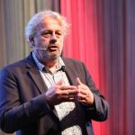 Ein Mann im Anzug spricht auf der Bühne mit einem Headset-Mikrofon und gestikuliert mit den Händen; hinter ihm rote und graue Vorhänge.