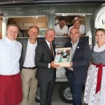 Sechs Personen posieren vor einem Imbisswagen; zwei Männer halten einen Geschenkkorb mit Flaschen und Lebensmitteln.