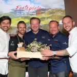 Fünf lächelnde Köche halten bei einer kulinarischen Veranstaltung vor österreichischer Bergkulisse eine Kiste mit Kräutern zusammen.