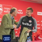 Zwei Männer schütteln sich bei einer Presseveranstaltung von Red Bull Salzburg die Hände und lächeln, während ein weiterer Mann in der Nähe sitzt.