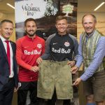 Vier Männer posieren in einer Halle, zwei von ihnen mit traditionellen Lederhosen, im Hintergrund Red Bull- und Salzburger Land-Fahnen.