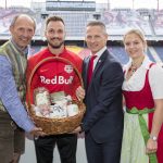 Vier Personen überreichen einen Geschenkkorb in einem Stadion; zwei tragen traditionelle österreichische Kleidung, einer einen Anzug, einer ein Red Bull Trikot.