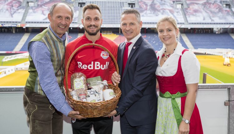 Vier lächelnde Menschen mit einem Geschenkkorb in einem Stadion, zwei in traditioneller Kleidung und zwei in moderner Kleidung.