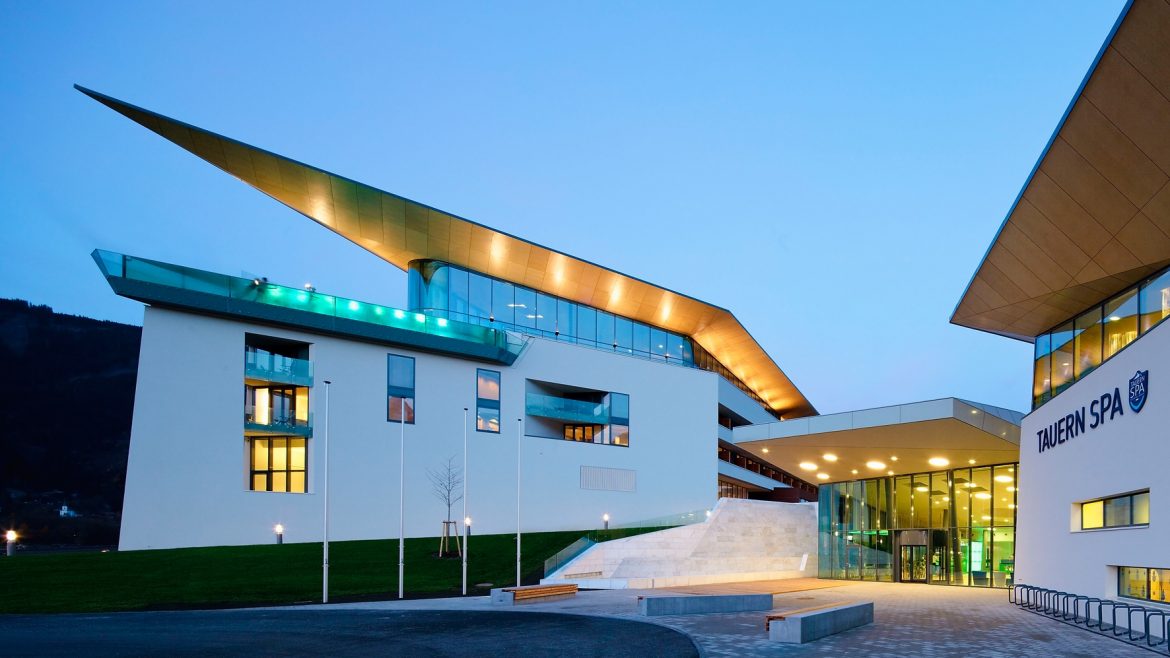 Modernes Gebäude mit kantigen Dächern und großen Fenstern, in der Dämmerung beleuchtet, Eingang mit der Aufschrift TAUERN SPA.